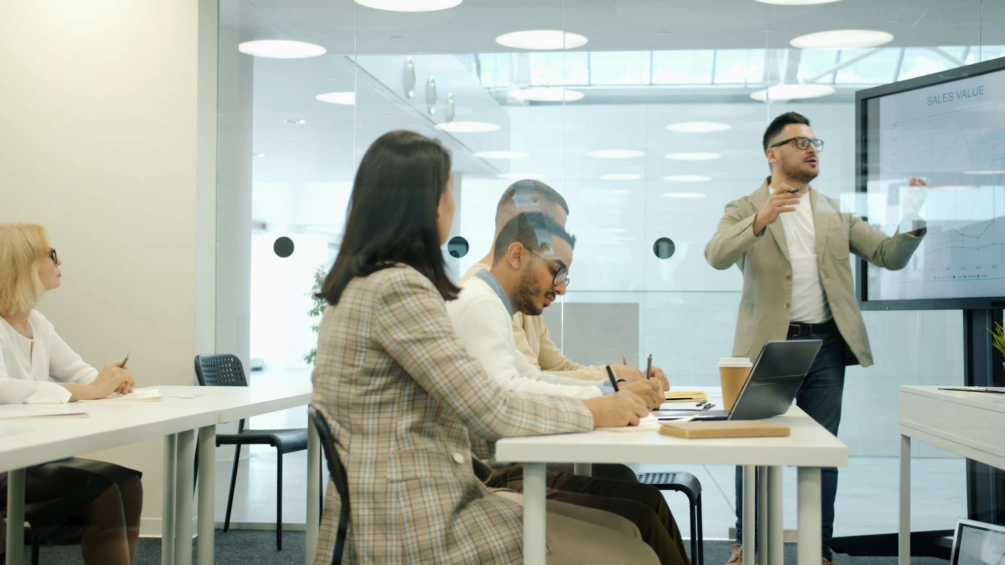 Instructor de pie junto a una pantalla con una gráfica de ventas mientras explica a cuatro personas de oficina sentadas en mesas, tomando notas en una sala luminosa de paredes de cristal.