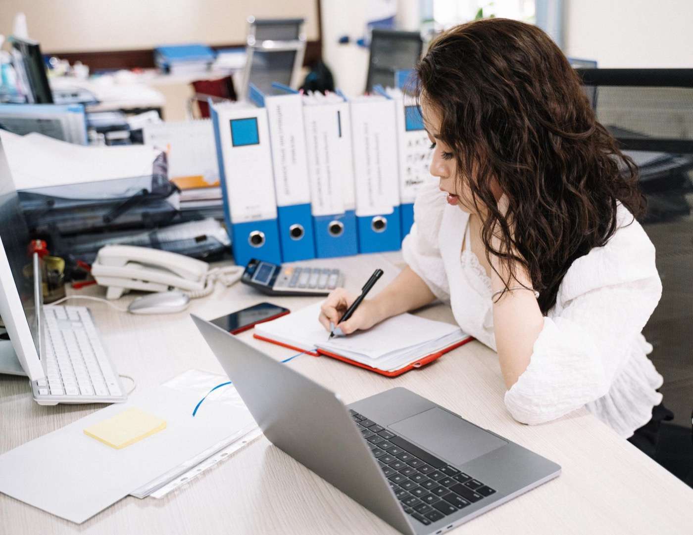Mujer de oficina escribe notas en una libreta frente a su laptop, en un escritorio con computadora de escritorio, teléfono fijo y carpetas azules de archivo.